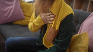 Sarı tişörtlü, göğsünü tutan kızıl sakallı bir adam kanepede rahat hissetmiyor..