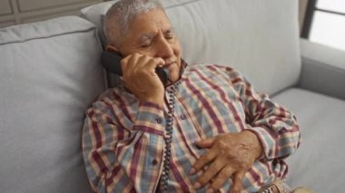 Yaşlı, beyaz saçlı İspanyol bir adam bir kanepede dinleniyor. Oturma odasında telefonla konuşuyor..
