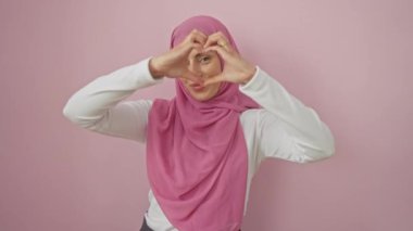 Tesettürlü neşeli, genç İspanyol kadın kalp şeklini ellerle şekillendiriyor, sevgi saçıyor. Soyutlanmış pembe bir arkaplan karşısında güzel bir portre. Romantik bir kavram..