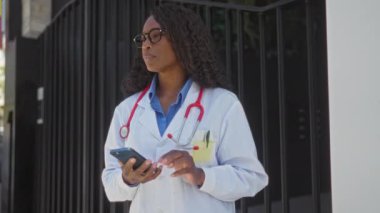 Kıvırcık saçlı Afro-Amerikalı bir kadın doktor şehir hastanesinin önünde akıllı telefon kullanıyor..
