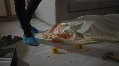 Eldivenli erkek bir adli tıp dedektifi, loş aydınlatılmış kapalı bir suç mahallinde kan lekeleri ve numaralandırılmış işaretlerle kanıtları inceliyor..
