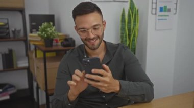 Modern ofis ortamında çalışırken sakallı, gülümseyen ve akıllı telefonunu kullanan yakışıklı bir İspanyol..