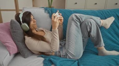 Akıllı telefonuyla video oyunu oynayan, kulaklık takan ve rahat bir oturma odasında kanepede dinlenen genç bir kadın..