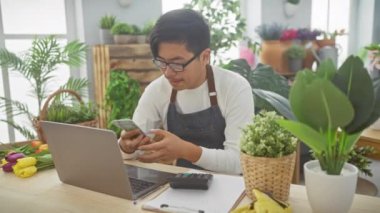 Asyalı bir adam yeşil bitkiler ve dizüstü bilgisayarla çiçekçide dolar sayıyor.,