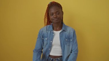 Kendine güvenen Afro-Amerikalı kadın kot gömlek giyiyor, izole edilmiş sarı arka planda duruyor, kameraya bakıyor, çene eliyle gülümsüyor, kollarını kavuşturup pozitif düşünce yayıyor..