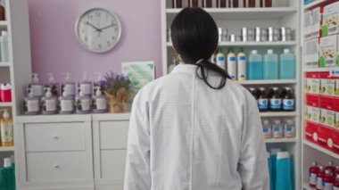 Beyaz laboratuvar önlüklü genç bir İspanyol kadının çeşitli ilaç ürünleri ve duvardaki saatle dolu parlak ışıklı bir eczanede durduğu görüldü..