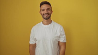Beyaz t-shirt giyen genç İspanyol adam ayakta ve baş parmağıyla yan tarafı işaret ediyor, izole edilmiş sarı arka planda ağzı açık gülümsüyor.