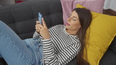 Genç bir İspanyol kadın evde rahatlar, renkli yastıklı bir kanepede uzanırken akıllı telefonunu karıştırır..