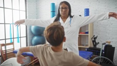 Bir kadın terapist, parlak bir fizyoterapi odasında egzersiz yapan genç bir çocuğa rehabilitasyon ekipmanlarıyla yardım ediyor..