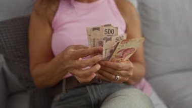 Evde Meksika pesolarını sayan bir kadın rahat bir iç mekanda el hareketlerini, para birimlerini ve bir oturma odasında günlük kıyafetleri vurguluyor..