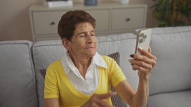 Evde rahat bir oturma odasında gri bir kanepede otururken akıllı telefon kullanan yaşlı İspanyol kadın..