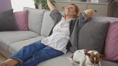 Kanepede evcil köpeğiyle rahat rahat oturan olgun bir kadın. Modern bir iç dekorasyonu olan rahat bir oturma odasında..