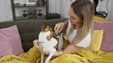 Genç bir kadın Jack Russell Terrier 'ını içeride rahat bir battaniyede tımar ediyor.