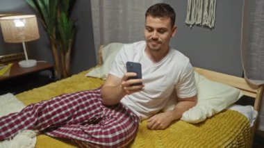 Pijamalı genç İspanyol bir adam rahat bir yatak odasında, sıcak bir ışık ve modern bir dekorun yer aldığı bir yatakta uzanırken akıllı telefonuyla bir video görüşmesi yapıyor..