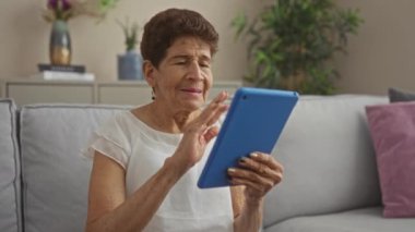 İç dekorasyon ve saksı bitkileriyle çevrili, rahat bir oturma odasında video görüşmesi sırasında mavi tablet kullanan yaşlı İspanyol kadın..