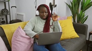 Evde dizüstü bilgisayar kullanırken kulaklıkla müzik dinleyen örgü ören Afrikalı Amerikalı kadın.