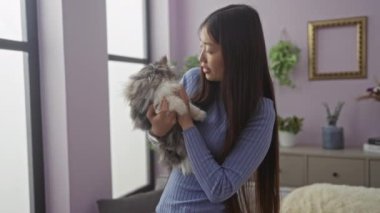 Konforlu bir oturma odasında genç Asyalı bir kadın ev bitkileri ve yumuşak ışıklandırmayla çevrili tüylü kedisini tutarak sıcak ve sade bir atmosfer yaratıyor..