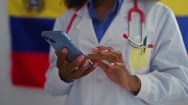 Afro-Amerikalı bir kadın doktor Venezuela bayrağının önünde steteskop ve laboratuvar önlüğü giymiş bir şekilde akıllı bir telefon tutuyor..