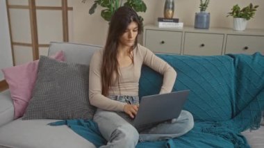 Oturma odasındaki kanepede dizüstü bilgisayarıyla oturan genç bir İspanyol kadın, işine odaklanmış ve dalmış bir şekilde 