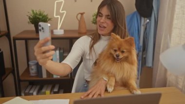 Genç bir kadın Pomeranian köpeğiyle evde selfie çekiyor, rahat bir odada neşe sergiliyor..