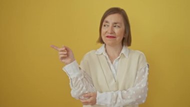 Neşeli orta yaşlı, İspanyol bir kadın mutlu bir gülümsemeyle ayakta duruyor, elini sarı bir arka plana doğru hareket ettiriyor.