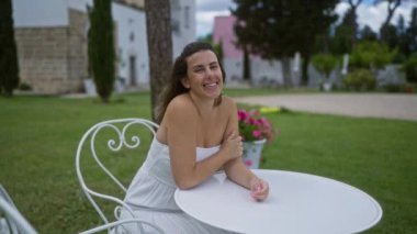 Beyaz elbiseli genç bir İspanyol kadın Puglia, İtalya 'da bir villada bir masada gülümsüyor ve oturuyor. Arka planda bir yaz günü çiçekler ve yeşillik var..