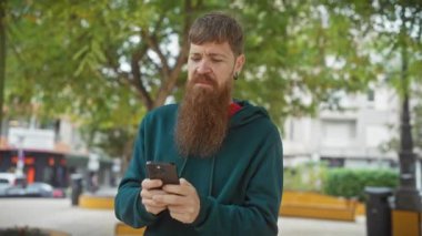 Sakallı kızıl saçlı adam parktaki akıllı telefona kaşlarını çatıyor..