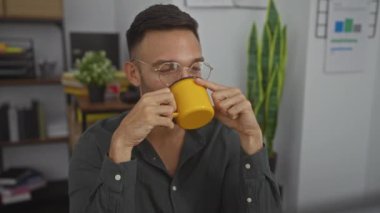 Sakallı ve gözlüklü genç bir İspanyol, arka planda bitki ve evrak işleri olan kapalı bir ofiste bir fincan kahvenin tadını çıkarıyor..