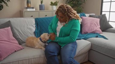 Örgülü genç siyahi bir kadın oturma odasında rahatlatıcı bir anın tadını çıkarıyor. Kanepede oturuyor ve rahat yastıklar ve dekorlar arasında evcil köpeğiyle etkileşime geçiyor..