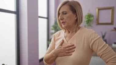 Evde oturan olgun bir kadın, göğsüne endişeyle dokunuyor, büyük pencerelerden gelen doğal ışık altında banyo yapıyor..