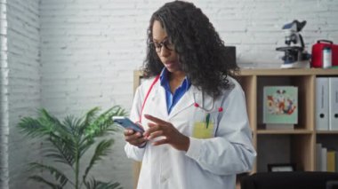 Beyaz önlüklü ve steteskoplu kıvırcık saçlı genç Afro-Amerikan kadın doktor bir klinikte çalışırken akıllı telefon kullanıyor..