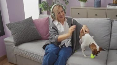 Olgun, sarışın, beyaz bir kadın oturma odasında oturmuş kulaklık takıyor ve gri bir kanepede pembe ve gri yastıklarla gülümserken köpeğini okşuyor..