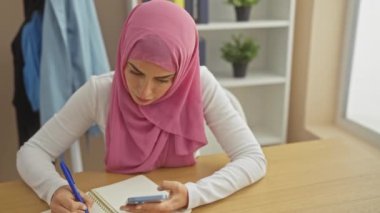Bir deftere yazı yazan ve rahat bir odada akıllı telefonunu kontrol eden tesettürlü Müslüman bir kadın..