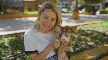 Güneşli bir günde, şehir parkında kahverengi bir chihuahua köpeği tutan gülümseyen sarışın kadın..