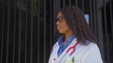 Afro-Amerikan kadın doktor bir şehir kliniğinin önünde tıbbi ceket ve steteskop giyerek duruyor kıvırcık saçlı ve gözlüklü özgüven ve profesyonelliği somutlaştırıyor..