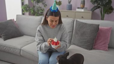 Parti şapkalı gülümseyen kadın içeride kanepede Chihuahua 'ya çilekli tatlı yediriyor.