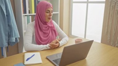 Mutlu, tesettürlü genç İspanyol kadın, bilgisayarını işaret ediyor, rahat evinde mutlu bir şekilde gülümsüyor..
