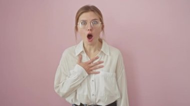 Şok olmuş ama heyecanlı genç sarışın kadın, gözlük takıyor, pembe arka planda duruyor, yüzü korku ve şaşkınlık ifadesi sergiliyor..