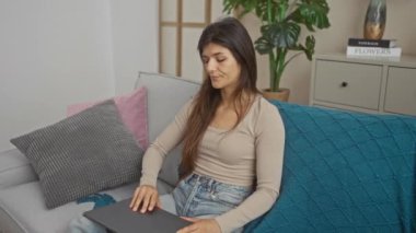 Genç esmer kadın oturma odasındaki kanepede oturuyor, dizüstü bilgisayar kullanıyor, ev dekorasyonuyla çevrili..