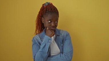 Endişeli bir Afrikalı Amerikalı kadın, kot gömlekli, sarı arka planda tek başına duruyor, endişeli görünüyor ve gergin bir şekilde bir soru üzerinde kafa yoruyor.