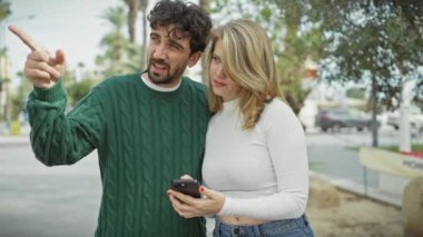 Bir adam ve bir kadın, güneşli bir şehir caddesinde, palmiye ağaçlarıyla kaplı bir akıllı telefonla iletişim kurarak birbirlerine yakınlık gösteriyorlar..