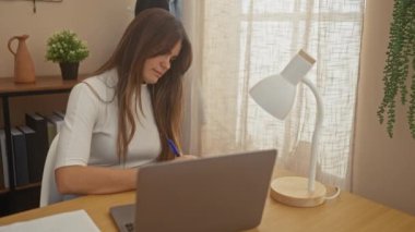 Odaklanmış genç bir kadın dizüstü bilgisayarda çalışıyor. Elinde lamba, bitki ve dekor var.