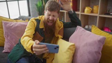 Sakallı yakışıklı kızıl adam kanepenin içinde selfie çekerken gülüyor.