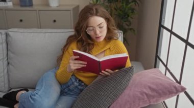 Gözlüklü ve sarı kazaklı genç bir kadın gri bir kanepede kitap okuyor. Yastıklı, çiçekli ve pencereli bir oturma odasında..