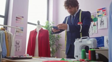 Yakışıklı adam terzi dükkanında manken, giysi, dikiş makinesi ve moda çizimleri ile akıllı telefon kullanıyor..