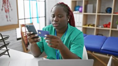 Akıllı telefon ve klinikteki kredi kartı bilgilerini inceleyen Afrikalı kadın sağlık uzmanı.