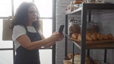 Ekmek ve pasta kavanozlarıyla dolu fırının içinde akıllı telefonla fotoğraf çeken, önlük ve gözlük takan, hamur işleri ve ekmeklerle rafların yanında duran bir kadın.