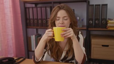 Genç ve çekici beyaz bir kadın, evindeki dosya ve kitaplarla çevrili sarı bir bardaktan su içiyor..