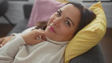 Orta yaşlı, gülümseyen İspanyol bir kadın, evde dinleniyor, sarı yastıklı bir kanepede, modern bir evde..