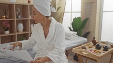 Kafasında havlu olan bir kadın elinde kağıt tutarken telefonla konuşuyor ve masaj masası ve yanında güzellik malzemeleri olan bir spa odasında oturuyor..